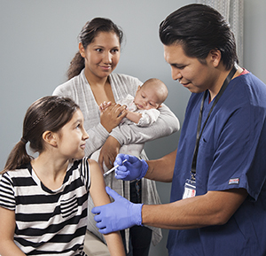 Healthcare provider giving girl injection in arm while woman with baby looks on.